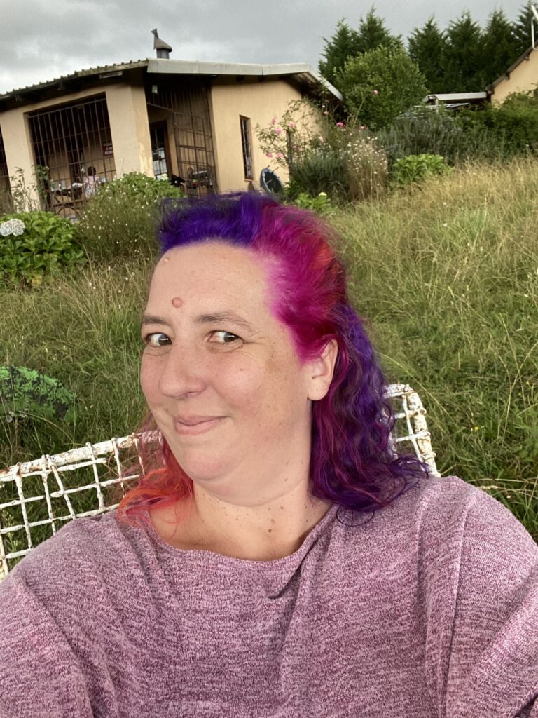 Photo of Bianca Johnson, Empowerment Coach, with rainbow hair sitting in her wild garden in front of her home in Rosetta. 